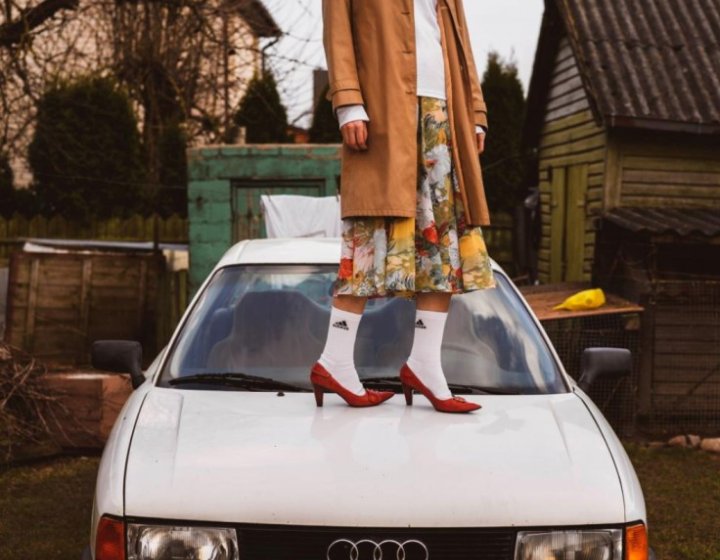 Agne stands on an old Audi, wearing white sports socks, red stilettos and an orange coat. 