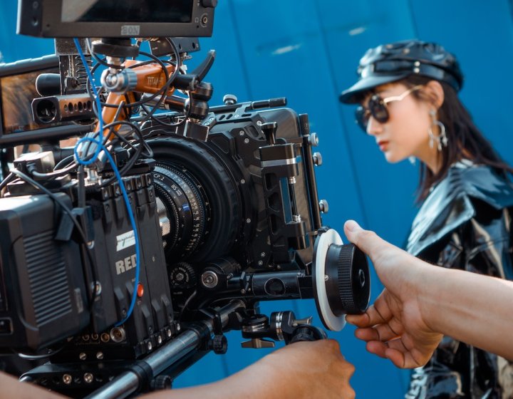 A man controlling a RED camera filming a women wearing sunglasses, a leather hat and jacket