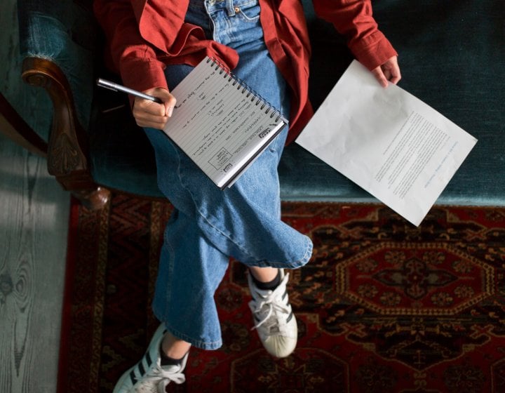 An aerial photo of a girl sat on a sofa with a notebook on her lap and a piece of paper next to her 