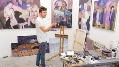 A male student standing at an easel surrounded by paintings