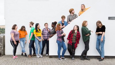13 Falmouth University students in different coloured tops stood against a white wall and on a white staircase.