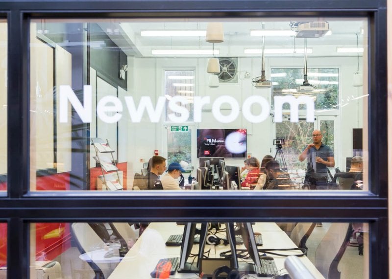 Through the window of the newsroom with lecturer talking to journalism students.