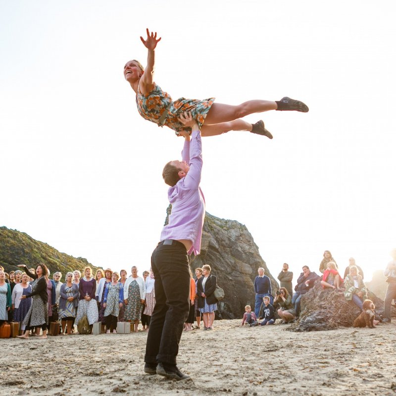 man raising a woman above his head on a beach surrounded by a crowd