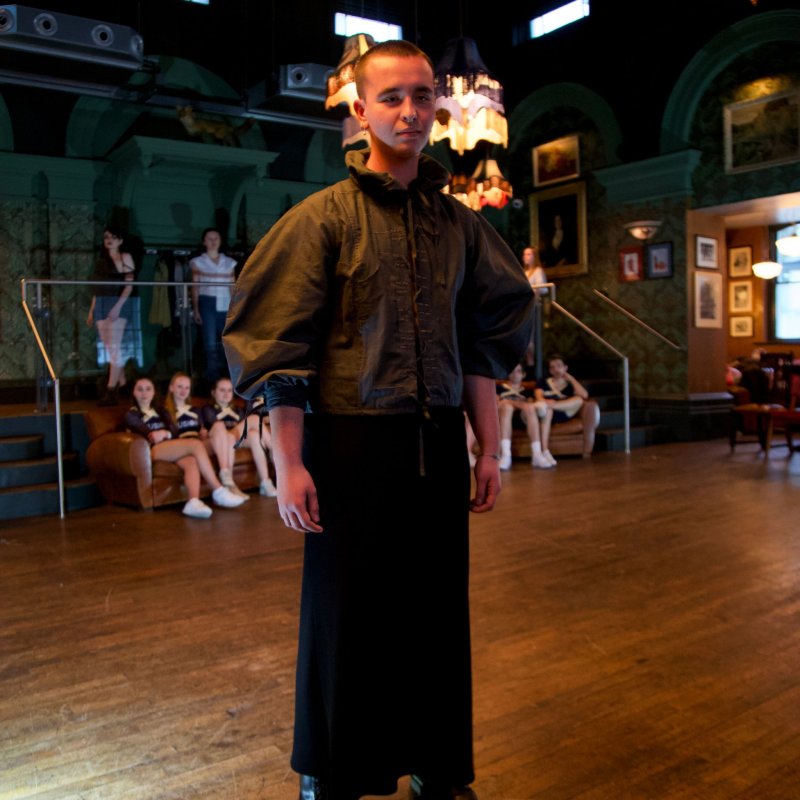 A man modelling on a catwalk. He wears black Dr Martens boots, black wide-legged trousers and an olive Tudor styled top. The top buffets at the arms, and the thick collar has been popped out by the model.