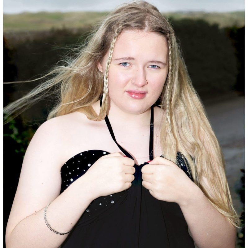 Portrait of Cornish girl wearing black dress, makeup and plaits in her hair against a countryside backdrop - from Cornish Maids photography project by Fran Rowse