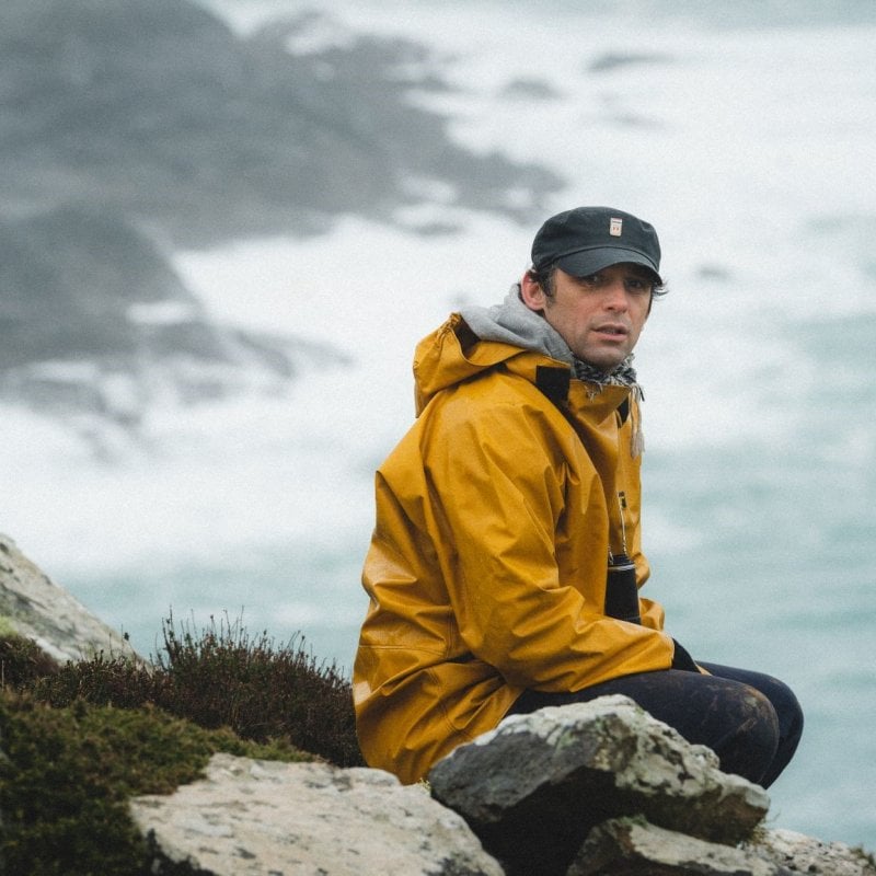 A man in a yellow coat sits atop a cliff, the ocean set behind him