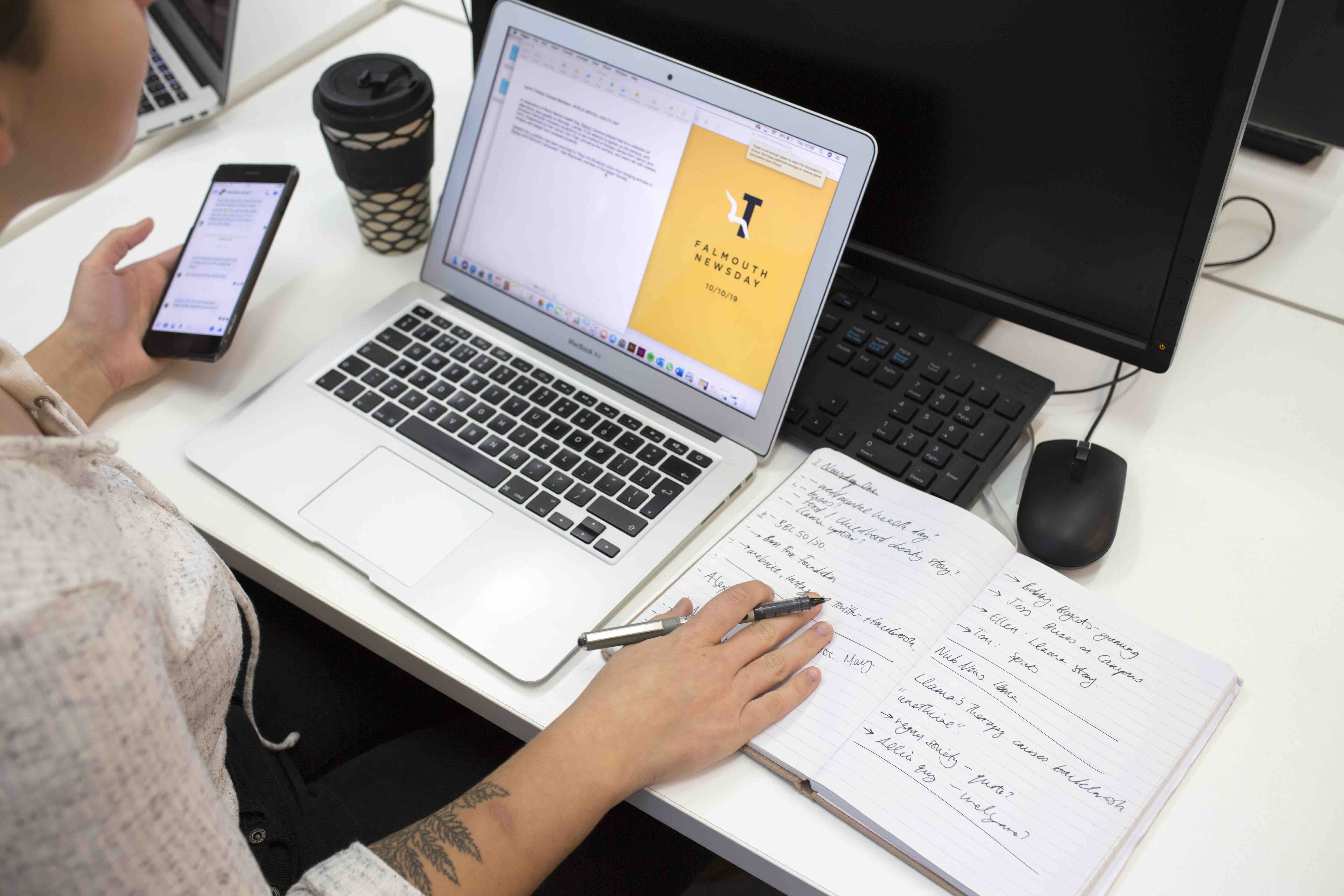 A Journalism student working at a laptop and holding a phone