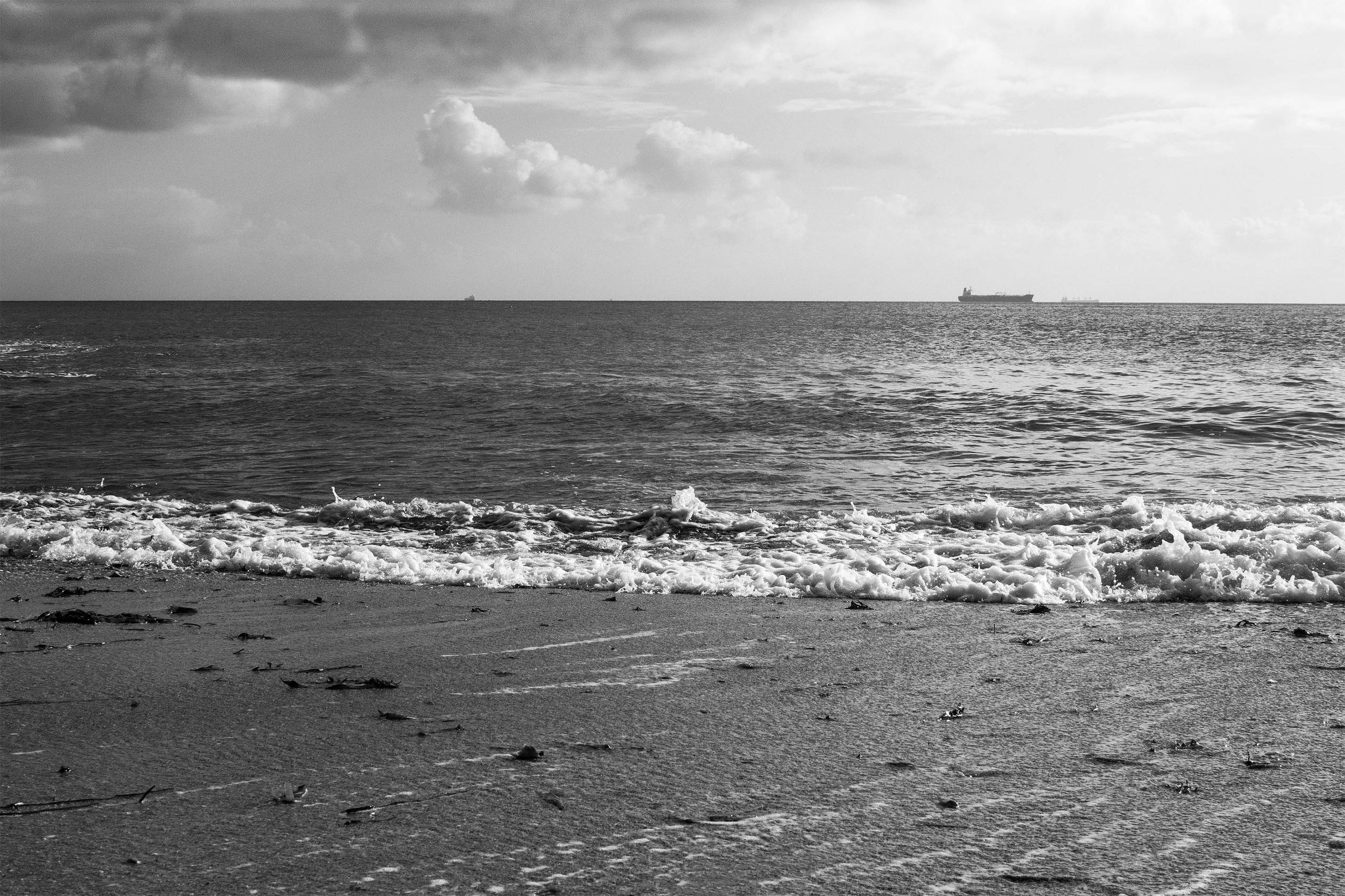 A black and white image of a beach, with foamy surf washing up against the sand. Two cargo ships can be seen in the distance