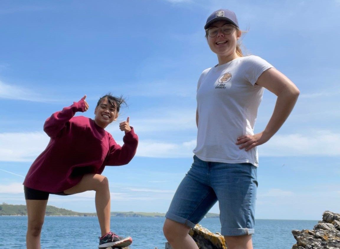 Two students posing for a photo on the beach with blue sky and sea in the background.