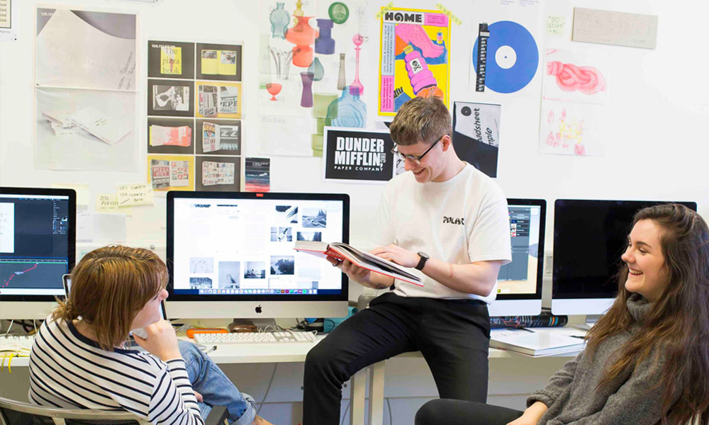Three Falmouth University students sat round an iMac with graphic design posters on the wall