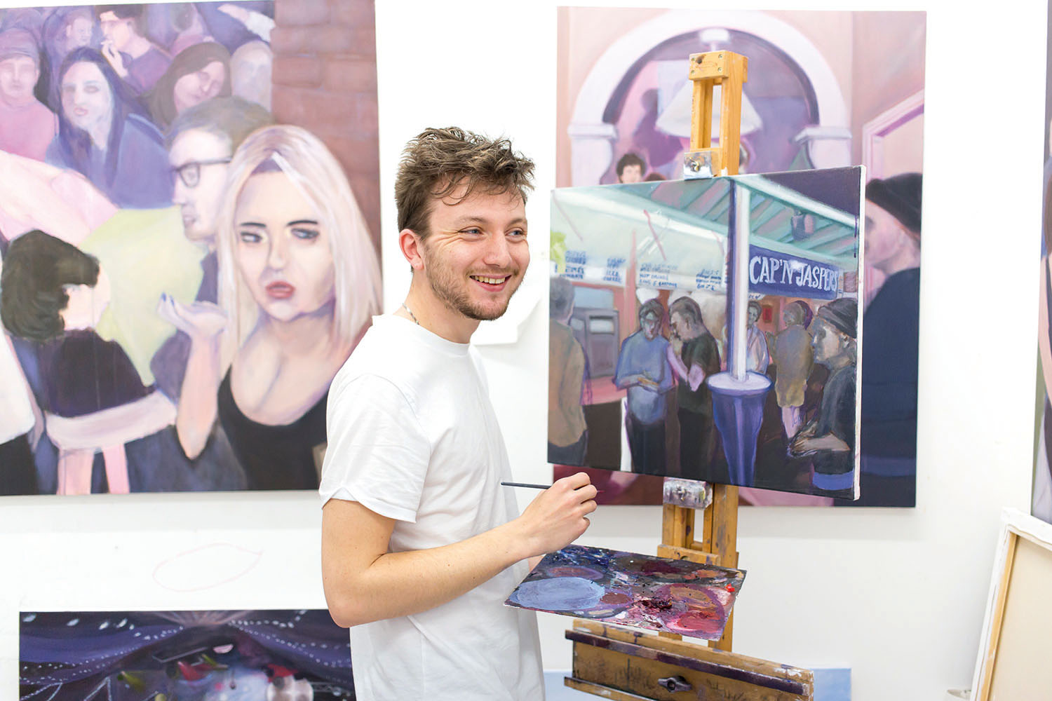 Student stood in front of paintings, smiling and holding paintbrush
