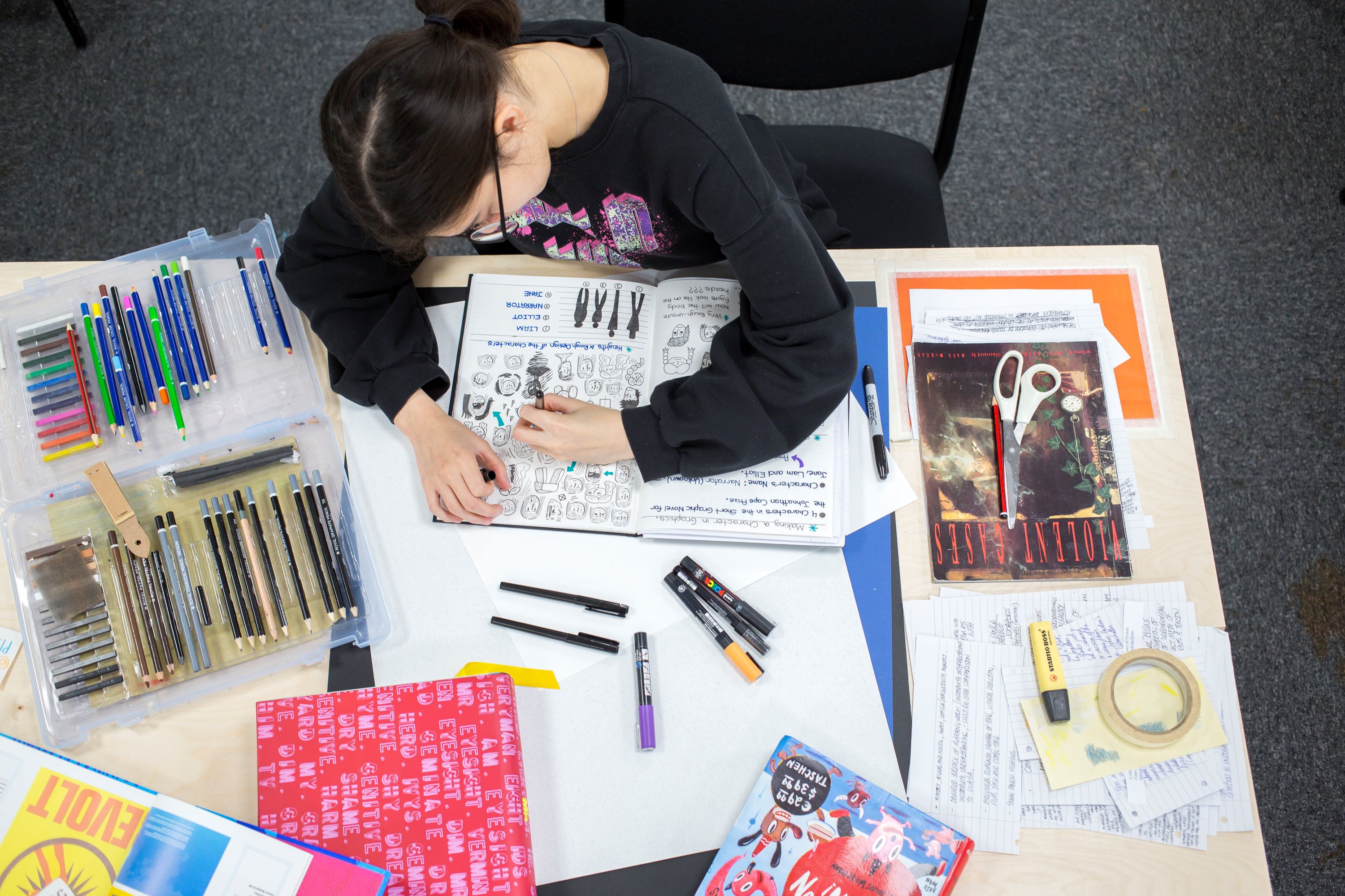 A female student drawing in a sketchbook surrounded by stationary