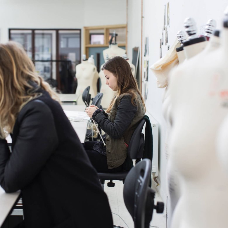 Two students in fashion studio, one on laptop and one knitting.