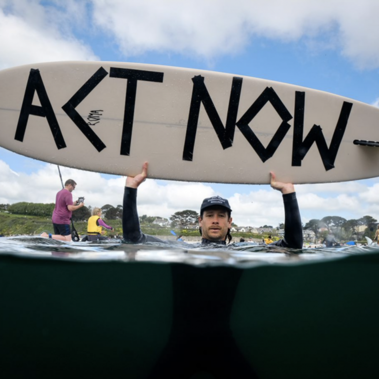 Activist in the sea holding up a surfboard with Act Now message written on it