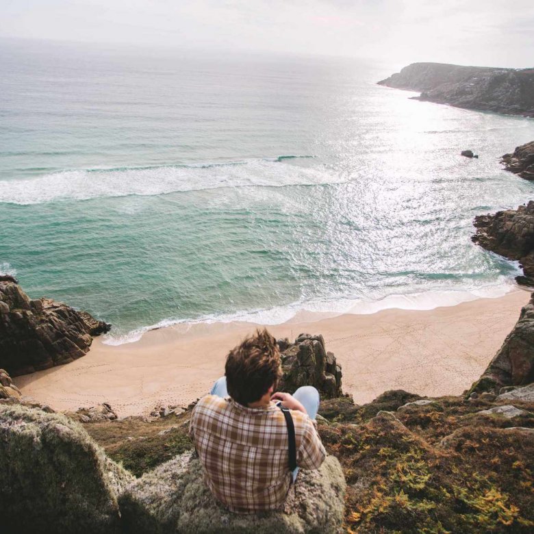 A Falmouth University student sitting on a cliff top overlooking a beach and sea