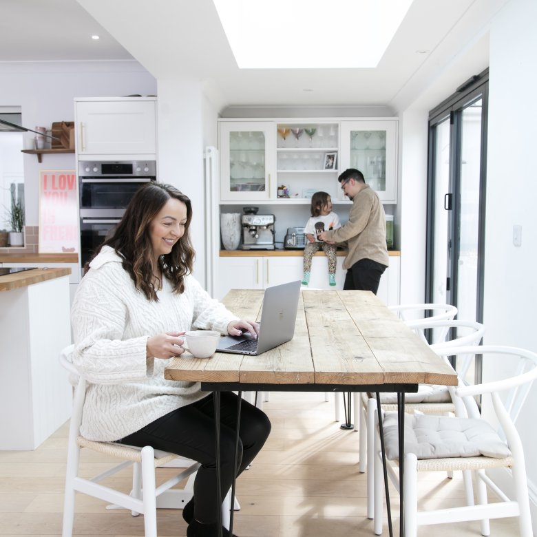 Woman sits at her dining room table with a laptop whilst her husband and child are in the background.