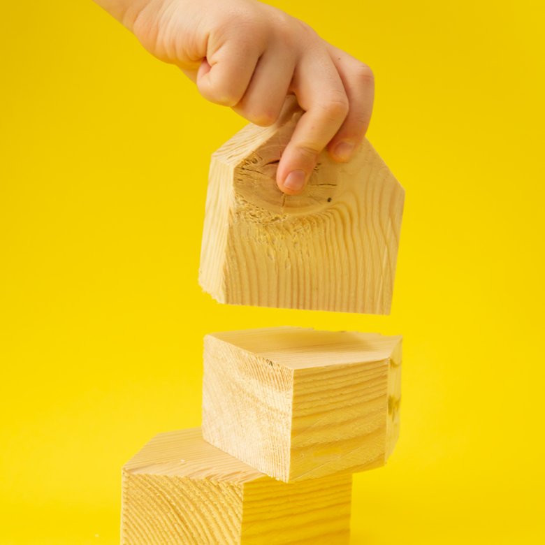 A set of three wooden blocks against a yellow background with a hand in shot holding one block 