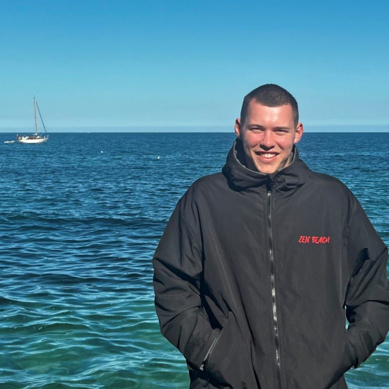 Portrait shot of Ben Penrose wearing black beach robe with red Zen Beach logo, with blue sea in the background