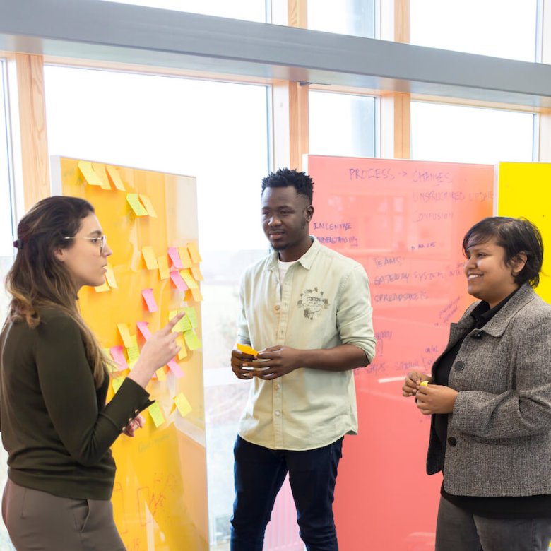 Three people standing in front of a yellow board covered in post-it notes