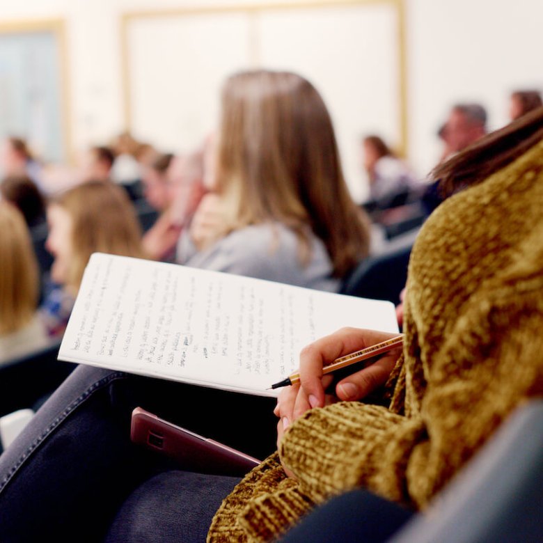 A person sitting in a lecture theatre with a notebook and pen in hand