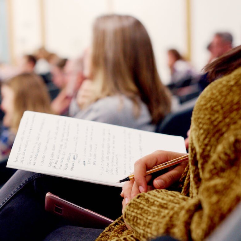 A person sitting in a lecture theatre with a notebook and pen in hand