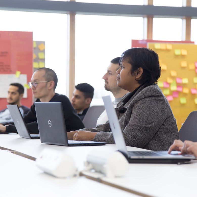 A group of people sat with laptops on a table, board of post-it notes behind them