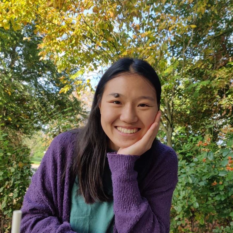 Female student wearing purple jumper 4smiling while sitting at wooden bench with leafy trees in the background.