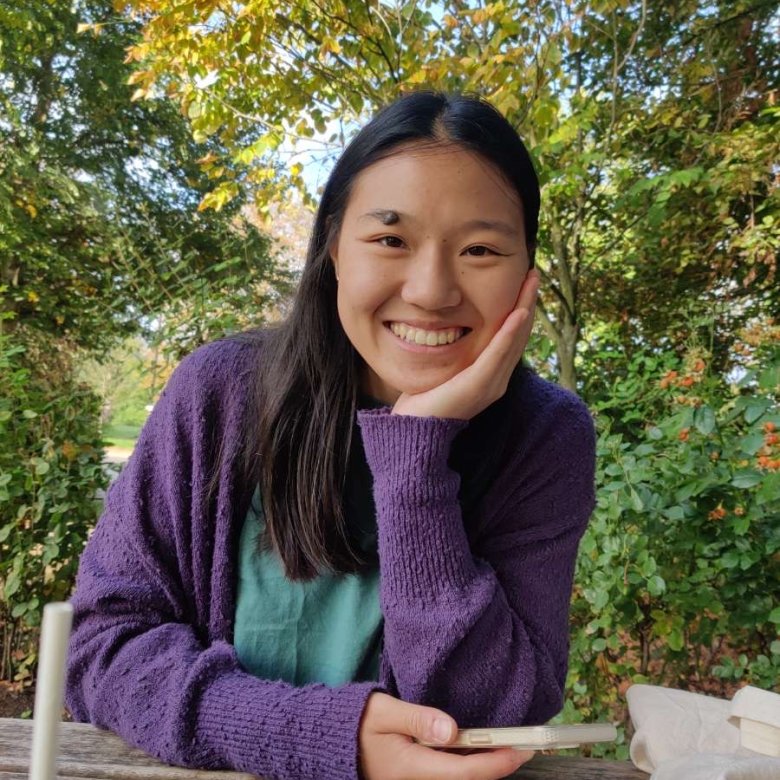Female student wearing purple jumper 4smiling while sitting at wooden bench with leafy trees in the background.