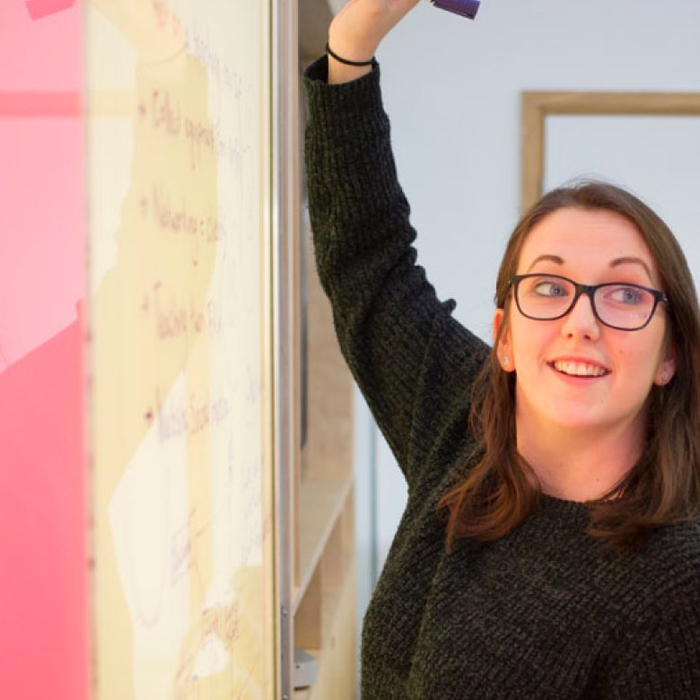 A woman wearing a black jumper, writing on a whiteboard with a pink panel