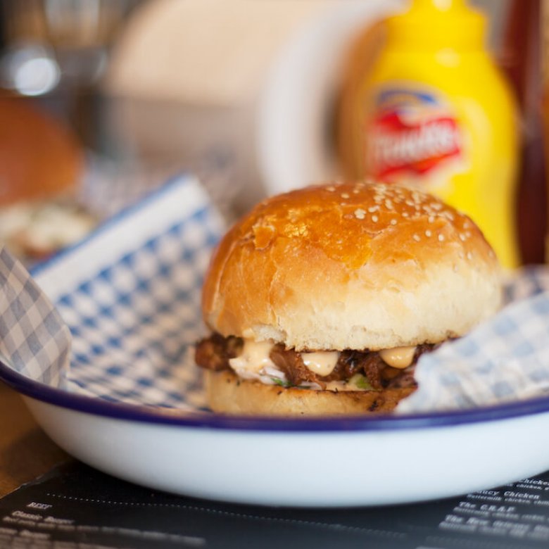 A burger in a white metal bowl with a bottle of mustard in the background