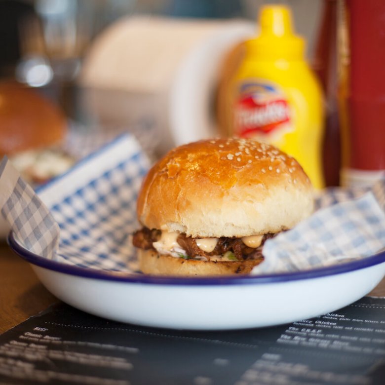 A burger in a white metal bowl with a bottle of mustard in the background