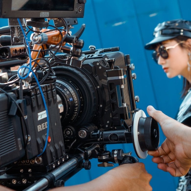 A man controlling a RED camera filming a women wearing sunglasses, a leather hat and jacket