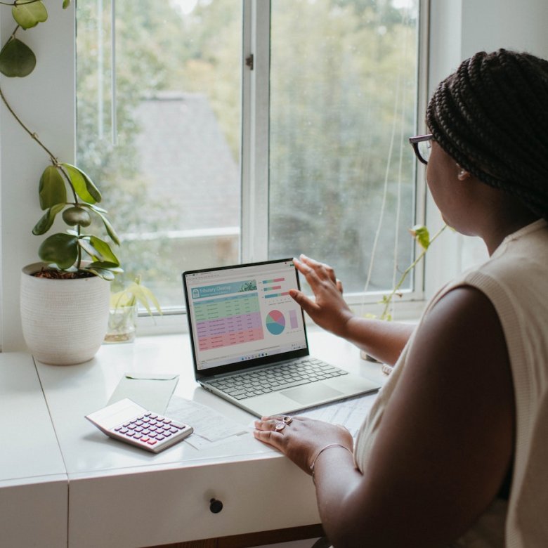 A woman working at a laptop by a window next to a plant and calculator