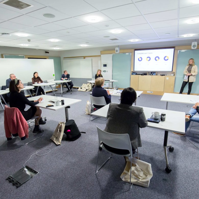 A student stands in front of a television screen and presents their business pitch to a panel of experts