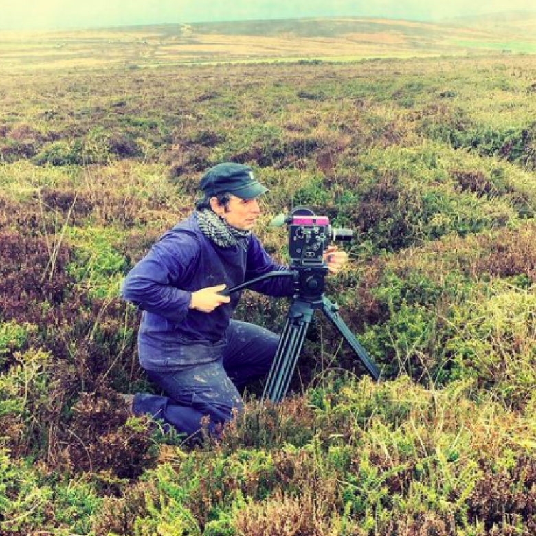 A filmmaker crouches behind his camera amongst shrubbery 