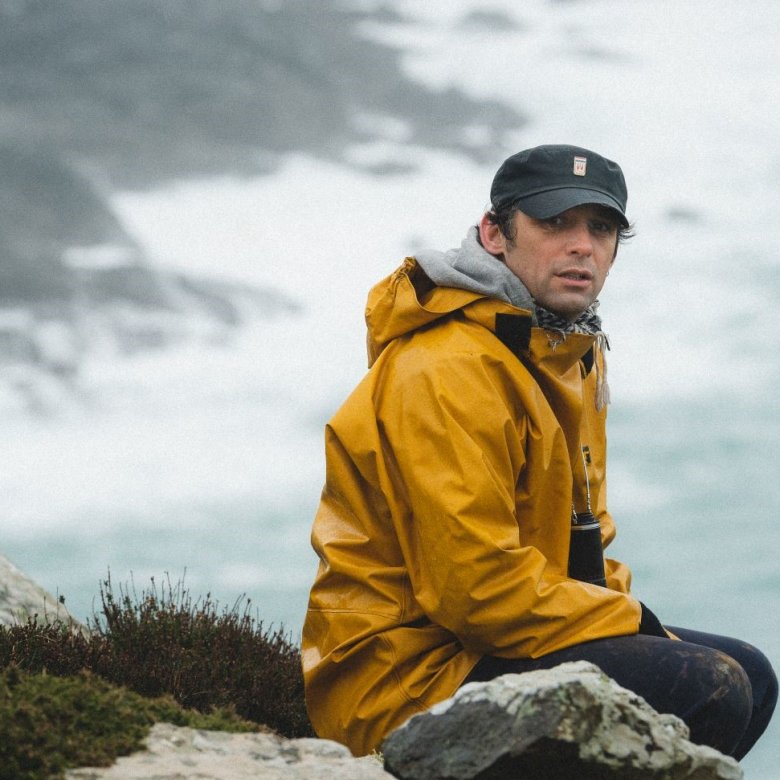 A man in a yellow coat sits atop a cliff, the ocean set behind him