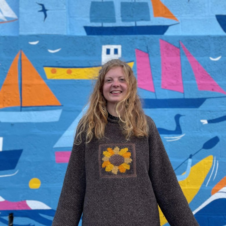 A girl wearing a brown jumper standing in front of a mural with boats
