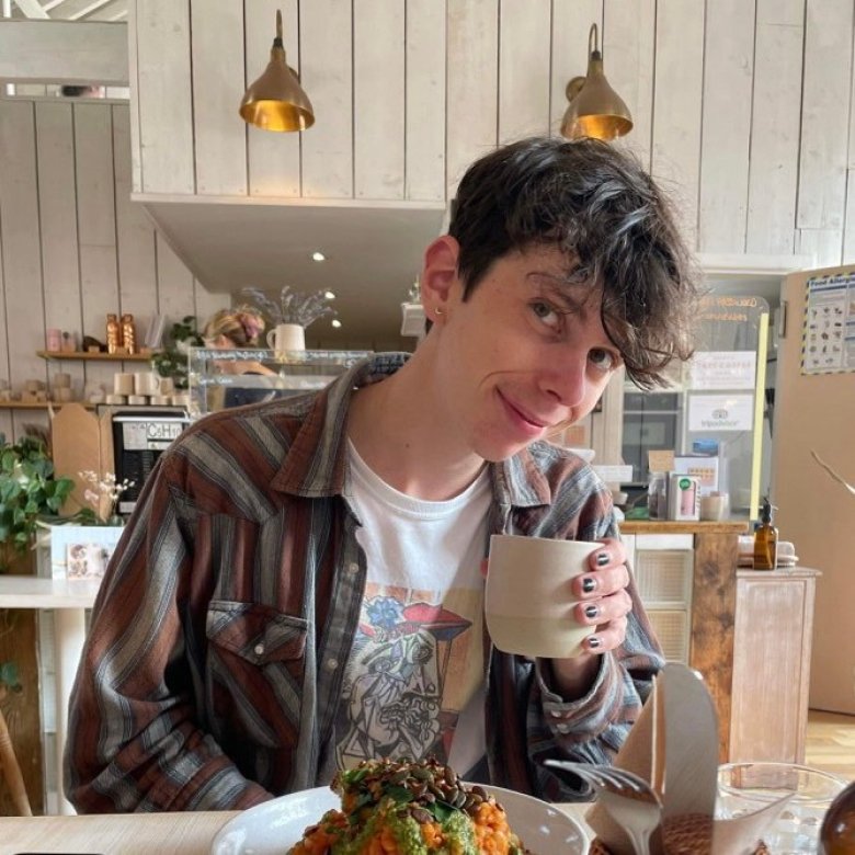 Photo of student Nathan Copeland wearing a graphic tee and checked shirt posing with a cup of coffee in a cafe.