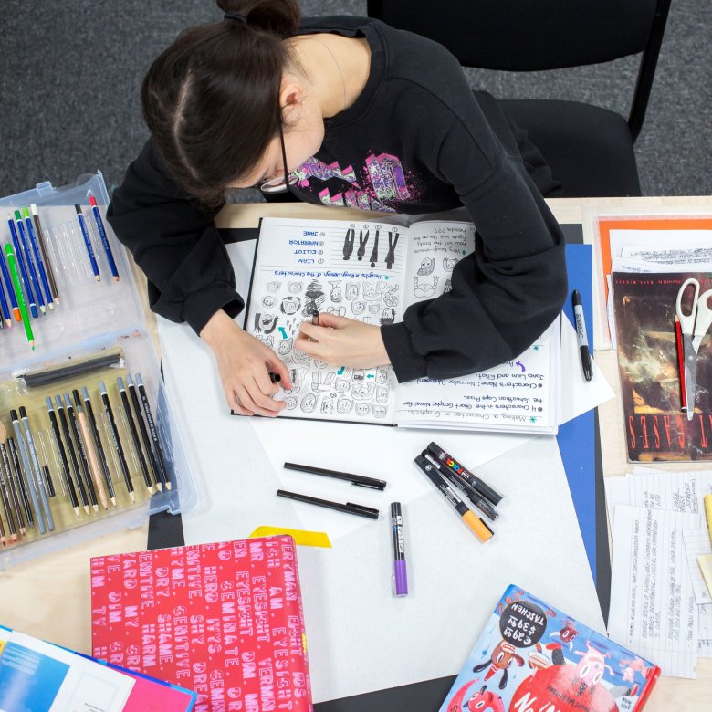 A female student drawing in a sketchbook surrounded by stationary