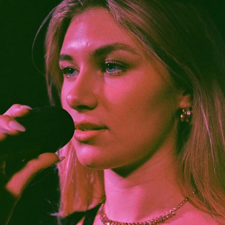 A close up of a woman singing into a microphone. She wears a gold necklace and is bathed in pink light