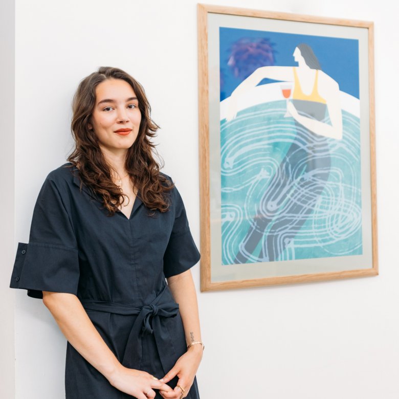 A young woman with dark hair standing next to a framed artwork of a woman drinking a glass of wine in a pool