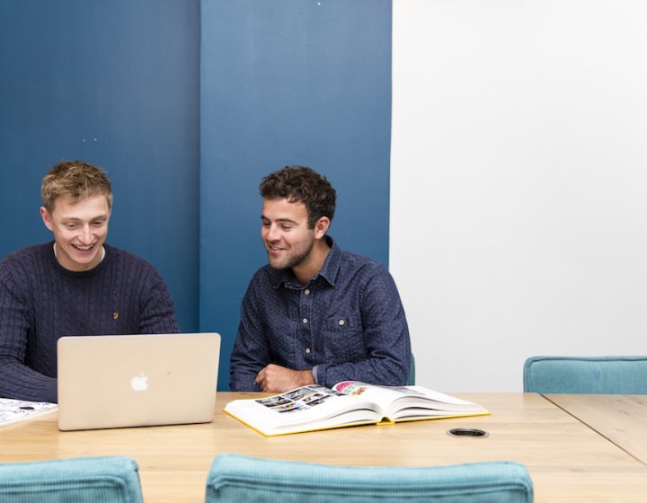 Two male students dressed in navy seated around a macbook 