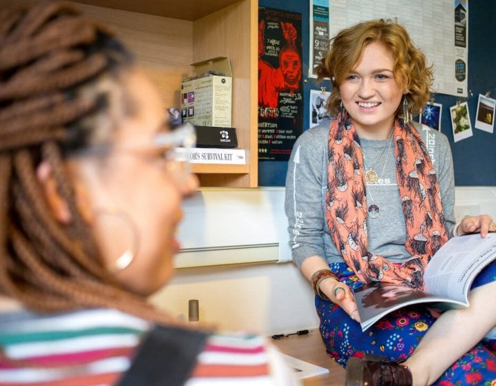 Student with book open on her lap chatting to another student in their room.