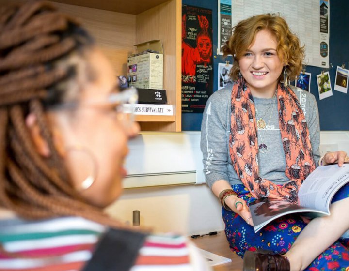 Student with book open on her lap chatting to another student in their room.