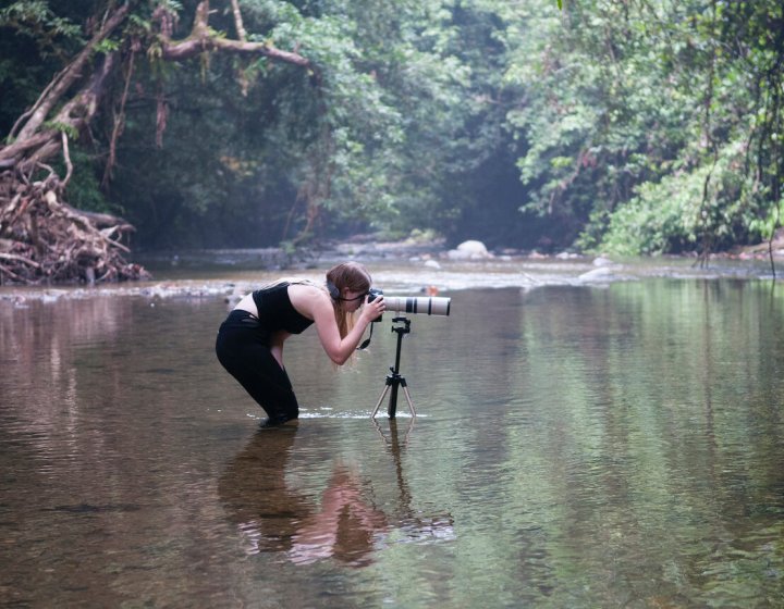 Falmouth University student immersed up to knees in river, bending down to view though camera on tripod.