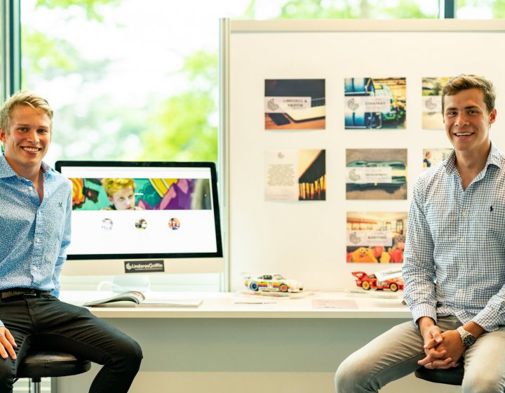 Falmouth University Business students in shirts sat in front of desk with work displayed.
