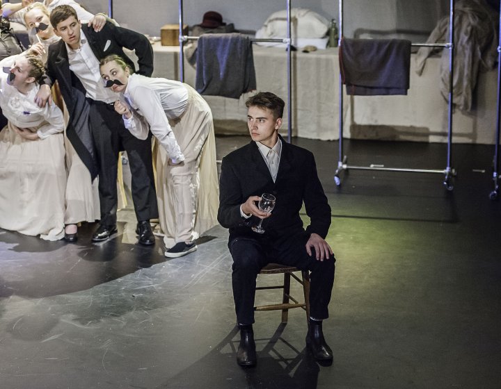 Actor in suit sat on chair on stage with wine glass in hand.