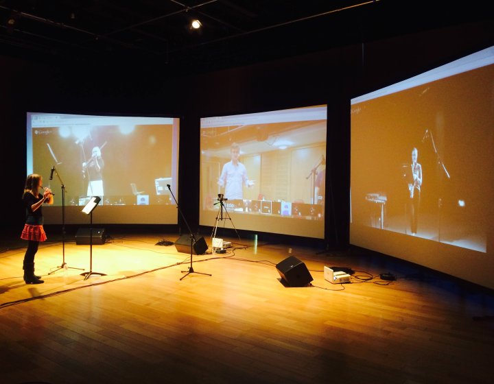 A woman playing the clarinet in front of three digital screens