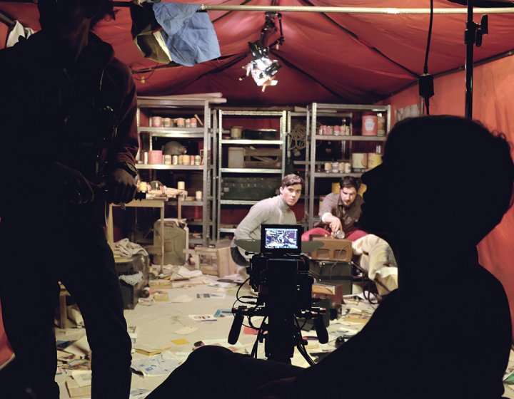 Silhouettes of crew on set in red tent, supplies on metal shelves in background, paperwork strewn across the floor.