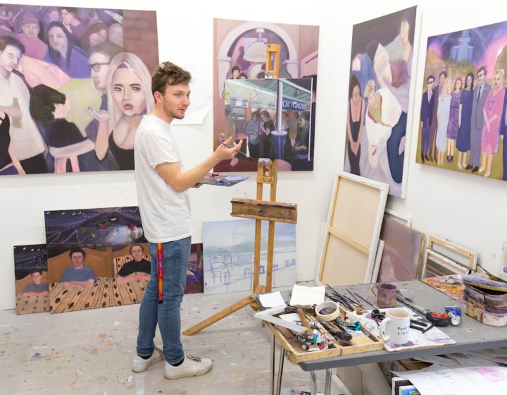 A male student standing at an easel surrounded by paintings